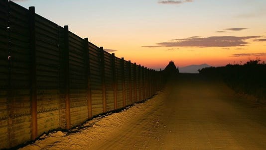 A US-Mexico border fence is illuminated by car headlights at sunset.